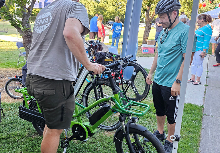 Two men look at an e-bike outside in a park.