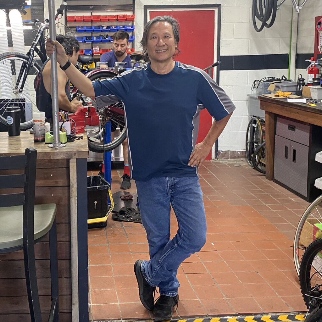 An Asian man stands in a bike shop with bikes behind him.