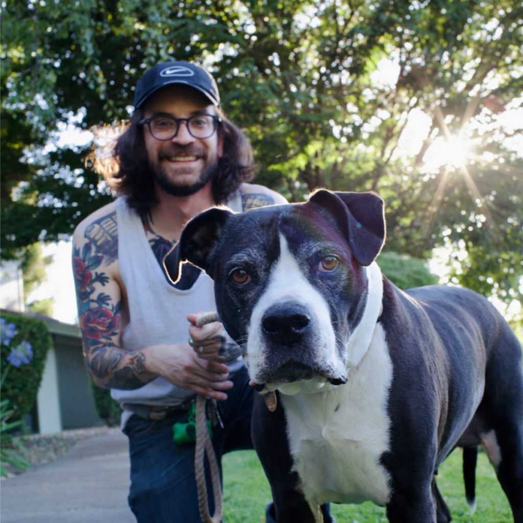 A man with long hair and tattoos kneels next to a medium sized dog.